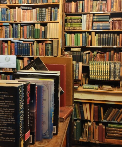 A dimly lit old book shop with floor to ceiling shelves. Books lie on the floors and worn carpets cover the aisles.