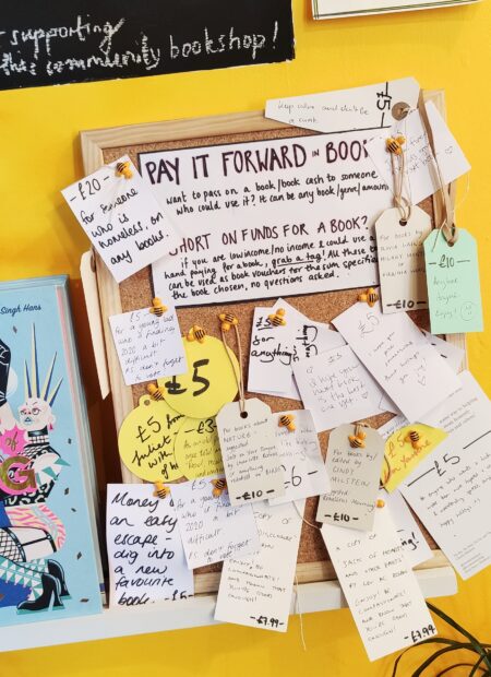 Pay it forward book pinboard at Lighthouse Bookshop. A brown corkboard against a yellow wall. Handwritten notices are stuck to it.