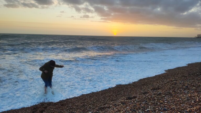 Alice is standing ankle-deep in the sea at brighton beach. A wave is splashing her legs.