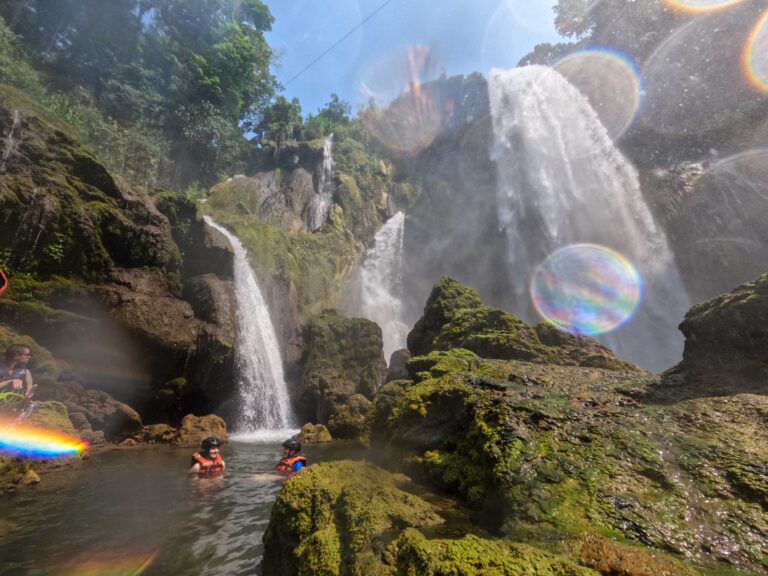 Pulhapanzak Waterfall surrounded by lush greenery. Two swimmers bathe in a pool