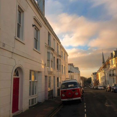 Terrace houses line the sides of a street. A vintage red campervan is parked outside a red door